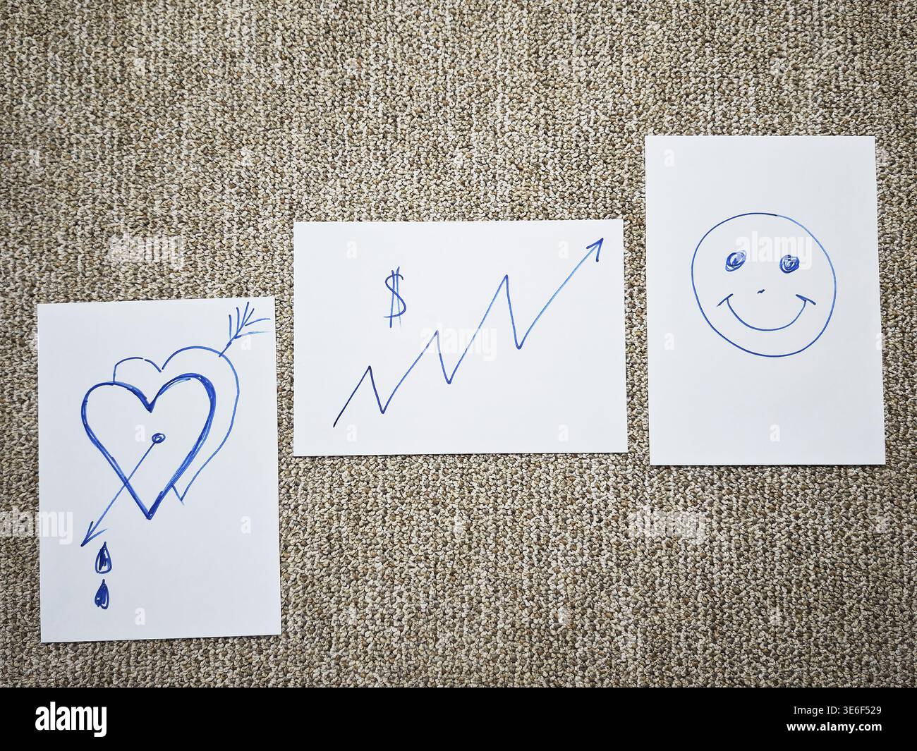 Tre disegni sono posizionati su una superficie grigia. I disegni mostrano un cuore spezzato, un simbolo del dollaro e una faccia sorridente che rappresenta sentimenti diversi. Foto Stock