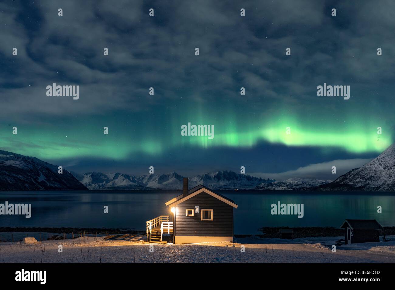 l'aurora boreale verde illumina il cielo invernale sopra un'accogliente cabina su una riva innevata vicino a Tromso, Norvegia, con calme acque del fiordo, montagne e basse nuvole Foto Stock