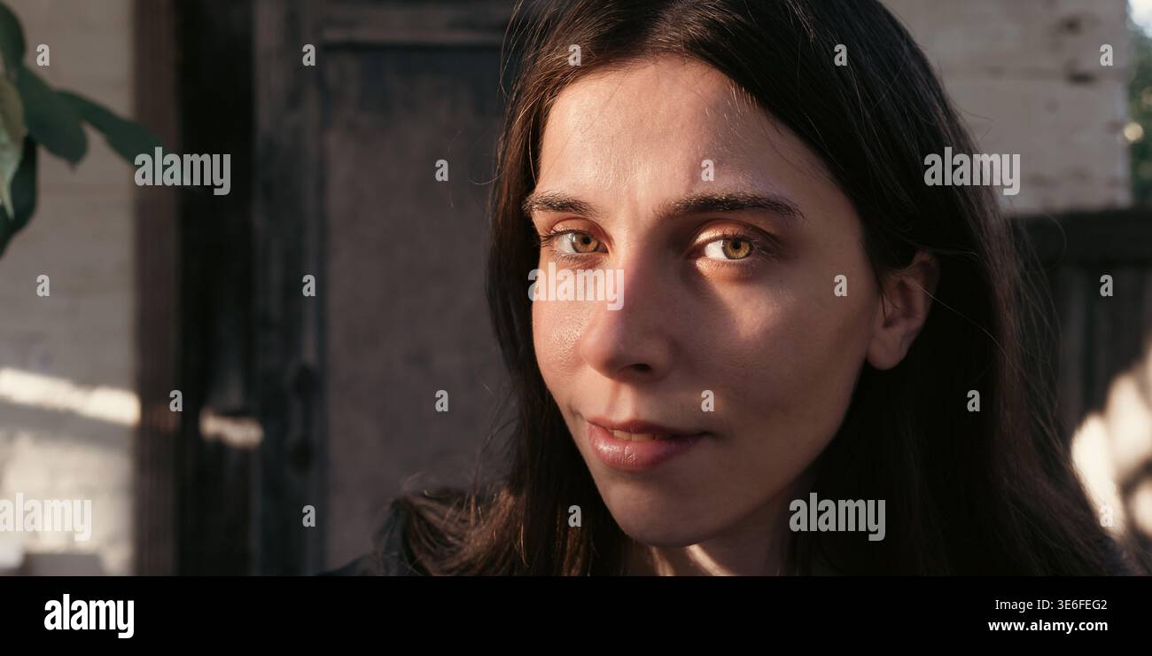 Chiudi il ritratto all'aperto di una giovane donna con un sorriso sottile in una calda luce naturale. Immagine di bellezza pulita con sguardo diretto, pelle sana e dorato morbido Foto Stock