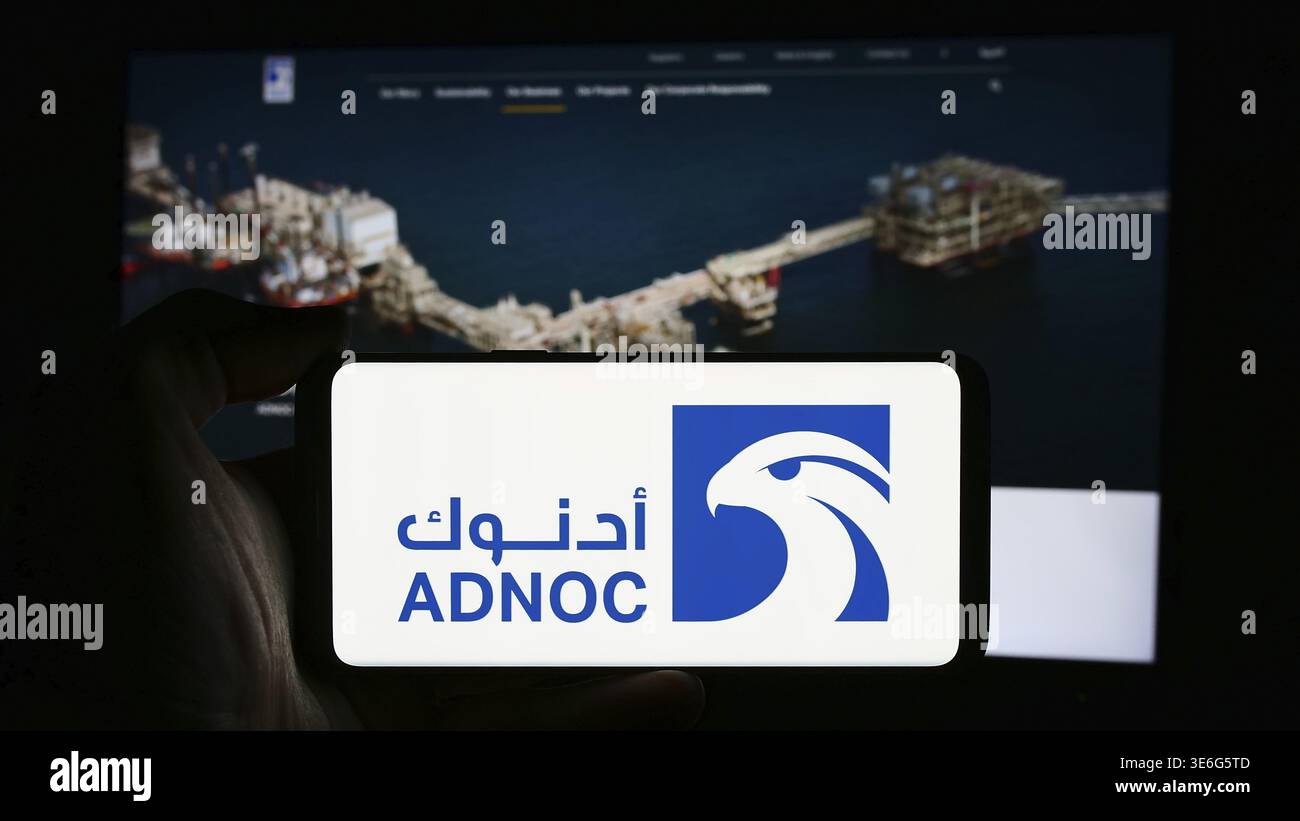 Stoccarda, Germania - 12-08-2024: Persona che detiene un cellulare con il logo dell'azienda petrolifera Abu Dhabi National Oil Company (ADNOC) di fronte alle pagine web aziendali Foto Stock