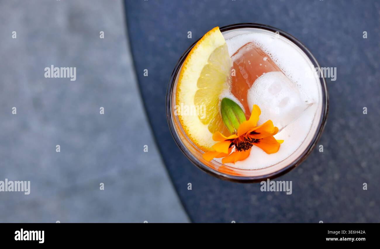 Primo piano un cocktail o un mocktail di colore arancione brillante in vetro alto, ricoperto di schiuma bianca, una fetta di agrumi e un fiore commestibile giallo fresco. Posizionato su un Foto Stock