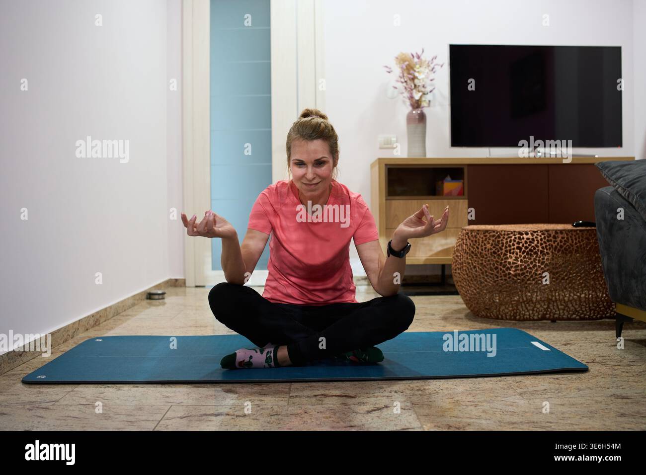 Donna di mezza età seduta a gambe incrociate in posa meditativa su un tappetino da yoga nel suo ufficio moderno Foto Stock