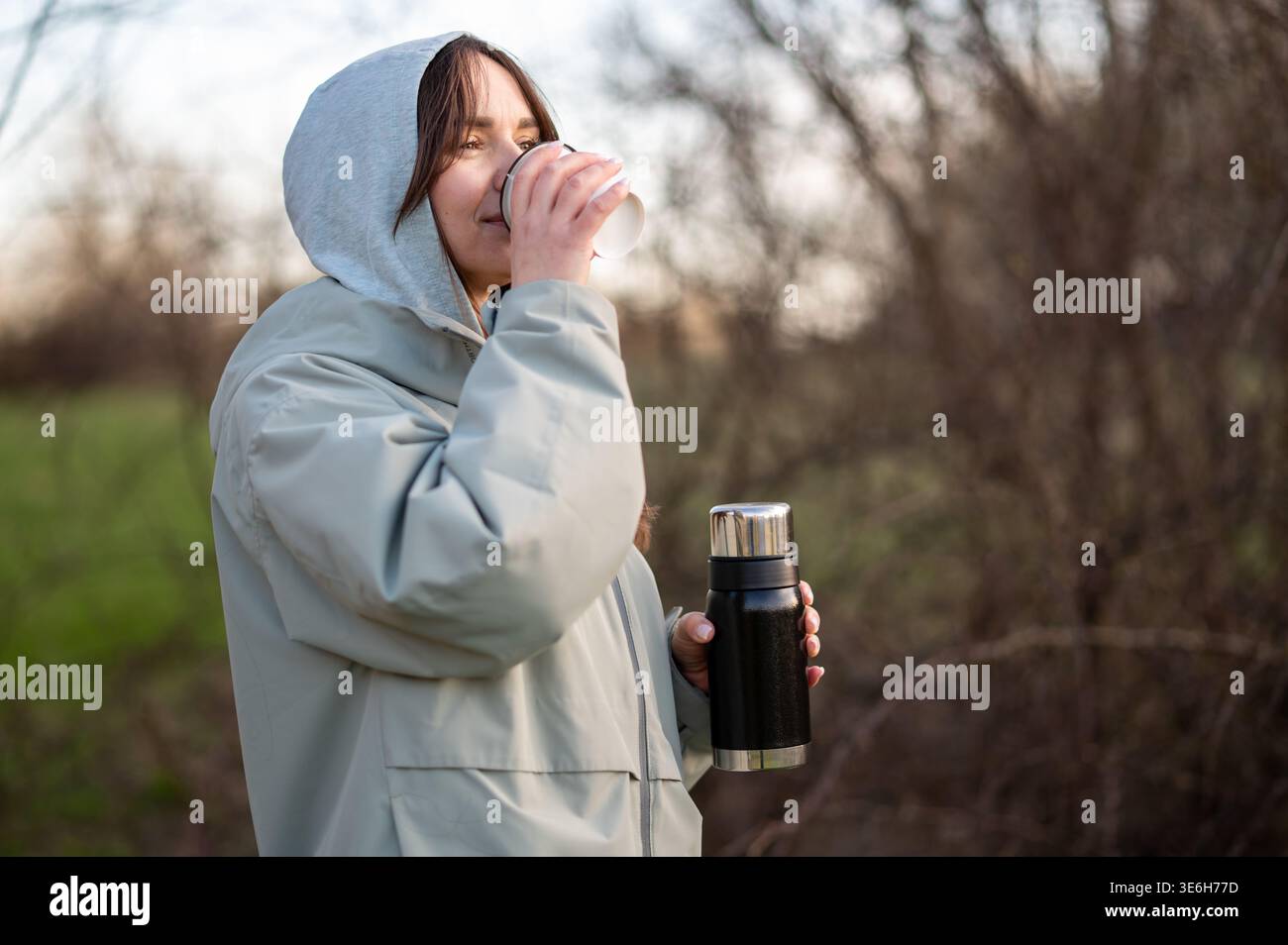 Donna sorridente in una giacca con cappuccio, mentre sorseggia un caffè o un tè all'aperto nel bosco durante una pausa. Persona con matraccio sottovuoto e bevanda in natura. Concetto di t Foto Stock