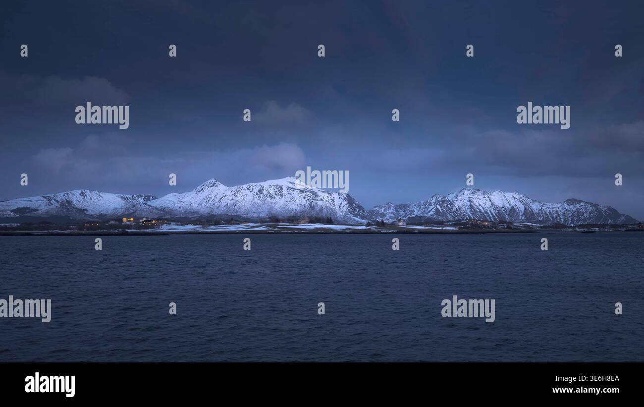 Una scena tranquilla si dispiega mentre il sole tramonta dietro le cime innevate di Leknes, Lofoten. Ombre morbide danzano sulle acque ghiacciate, creando un ambiente sereno Foto Stock