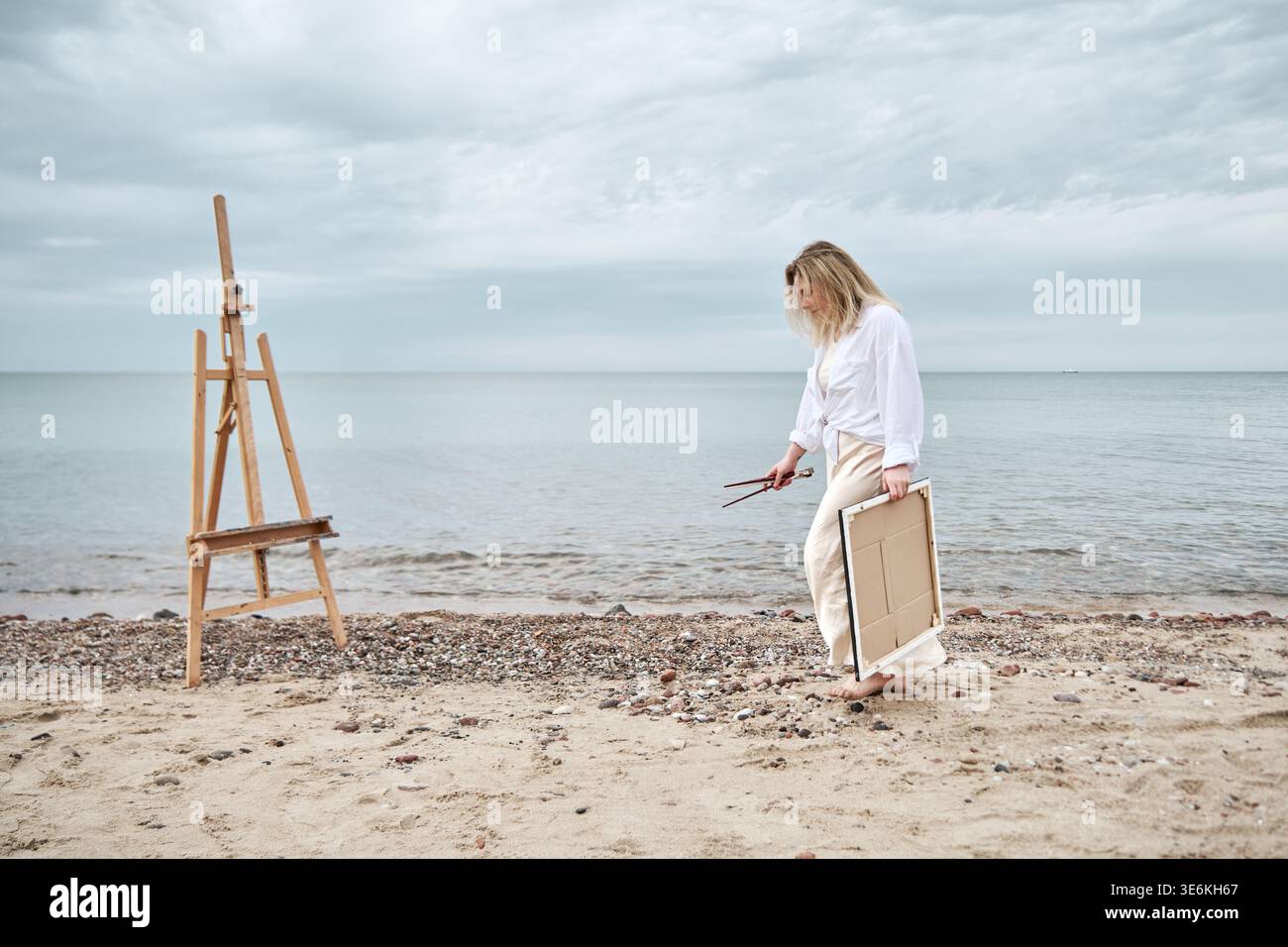 Una donna adulta con i capelli biondi porta tela e spazzole verso il cavalletto allestito sulla spiaggia sabbiosa sotto il cielo nuvoloso. Il mare calmo e i toni silenziosi creano un tranqui Foto Stock