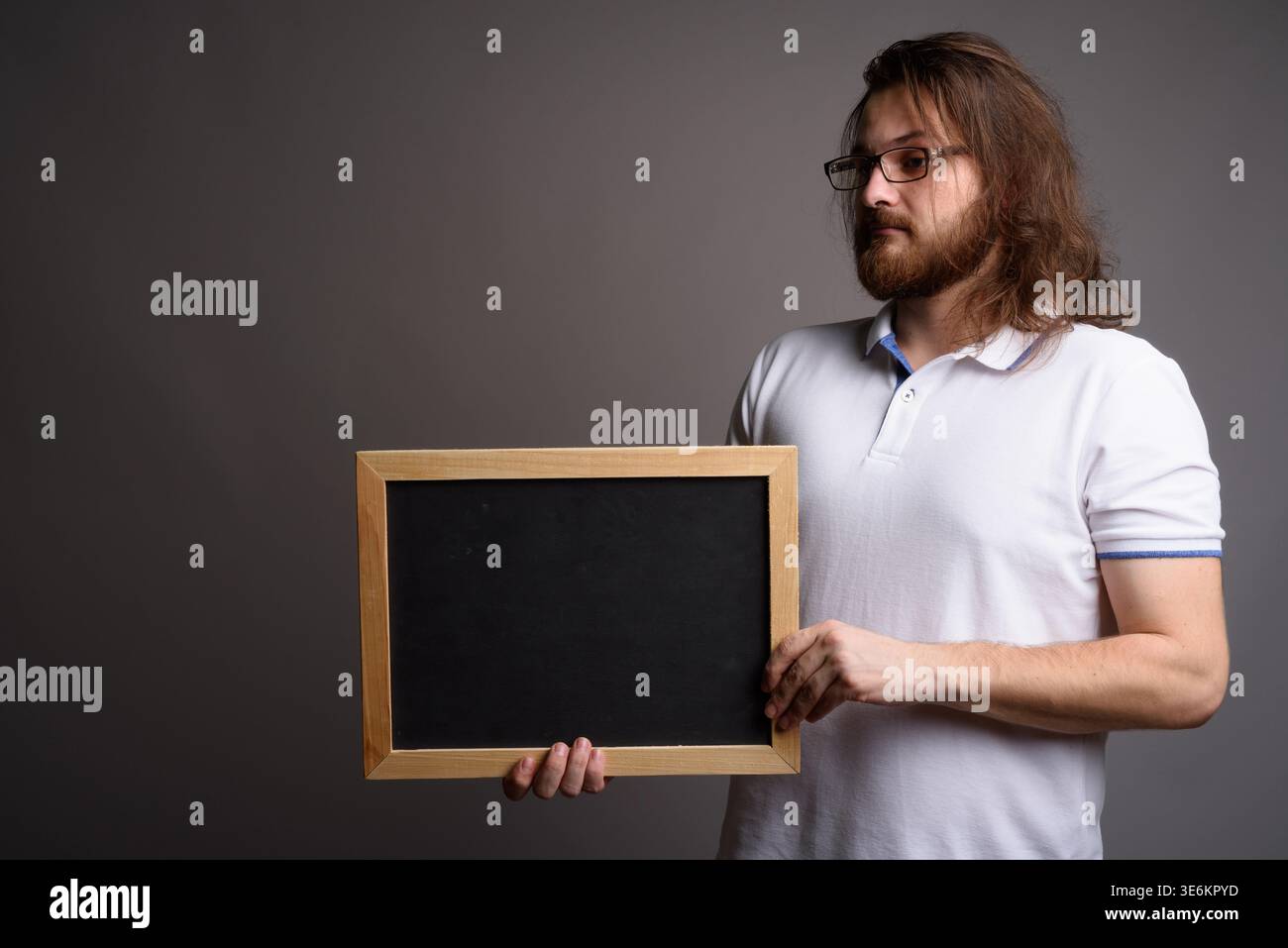 Ritratto in studio di un giovane e premuroso uomo caucasico con capelli lunghi che reggono una lavagna vuota. Ideale per messaggi personalizzati, idee visionarie e promozioni Foto Stock