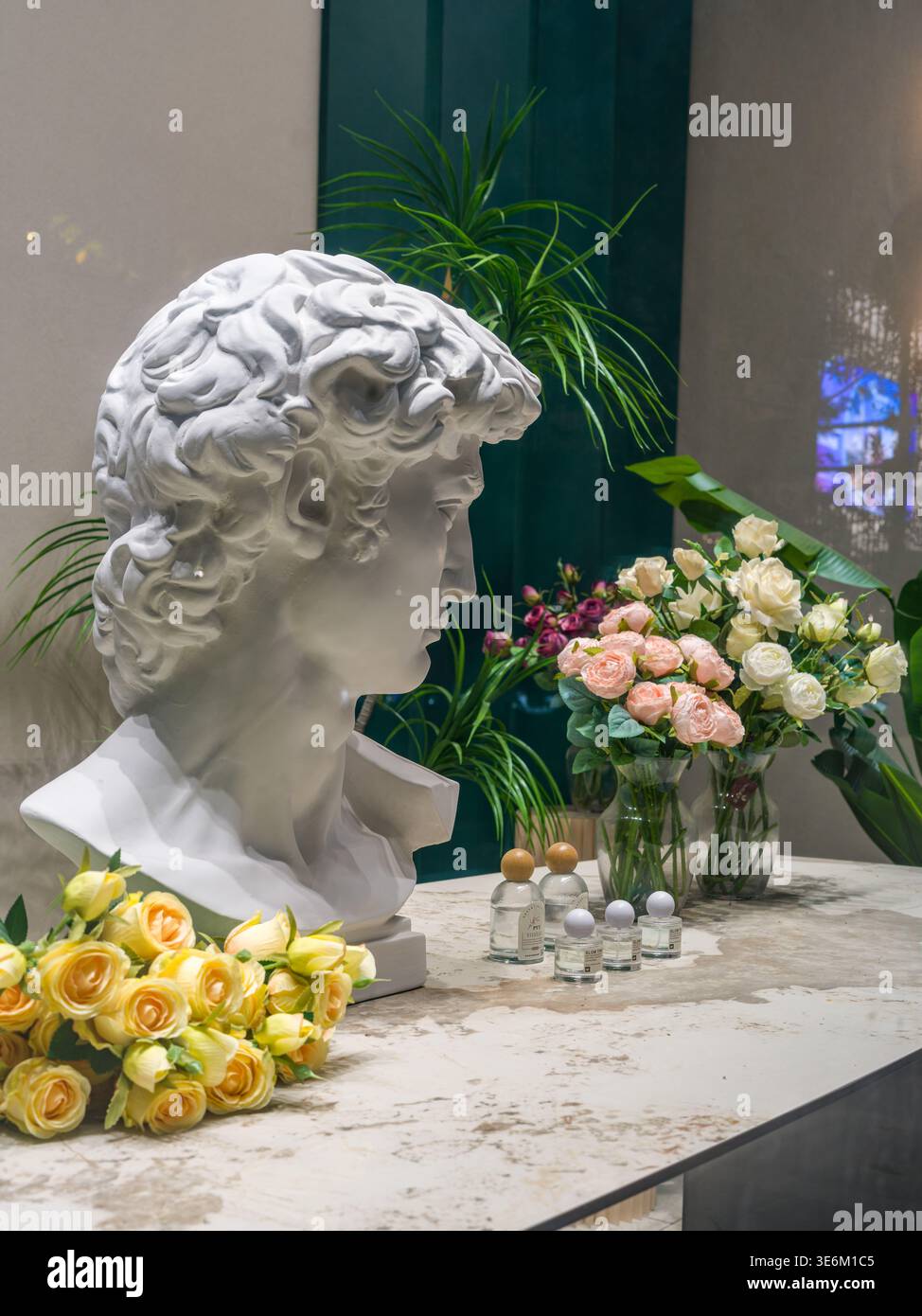 Elegante esposizione interna con una classica statua bianca del busto in gesso, un bouquet di rose gialle e rosa e piccole bottiglie di profumo su un marmo su Foto Stock