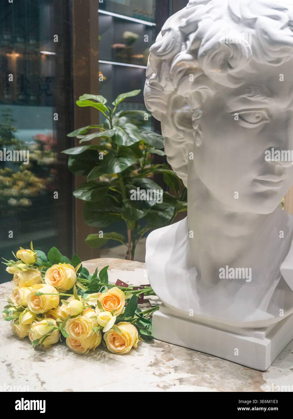Elegante esposizione interna caratterizzata da una classica scultura di busto in gesso bianco e un bouquet di rose gialle su una superficie in marmo. La scena rappresenta lu Foto Stock