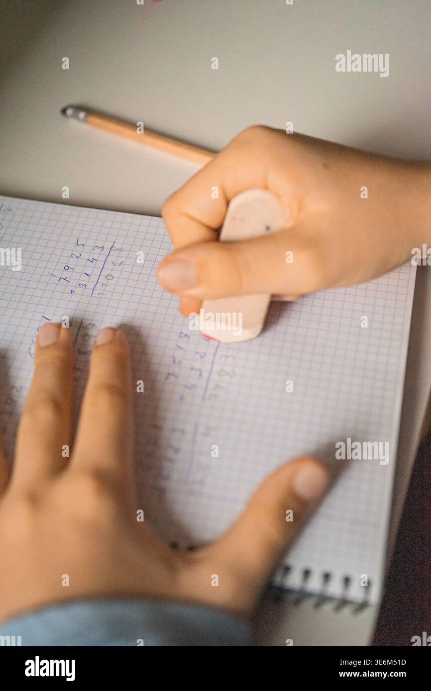 Primo piano delle mani di un bambino utilizzando una gomma per cancellare le operazioni matematiche scritte in un quaderno di carta grafica. Concetto di studio, apprendimento e istruzione di base Foto Stock