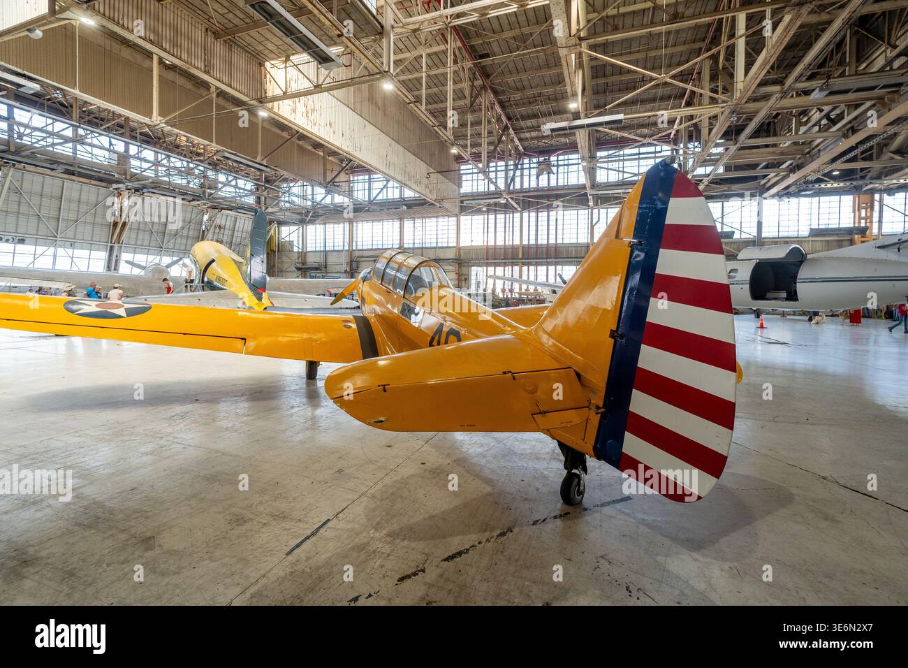 Un velivolo da addestramento giallo d'epoca con strisce di coda americane si trova in uno spazioso hangar, circondato da altri aerei storici e visitatori durante un avi Foto Stock