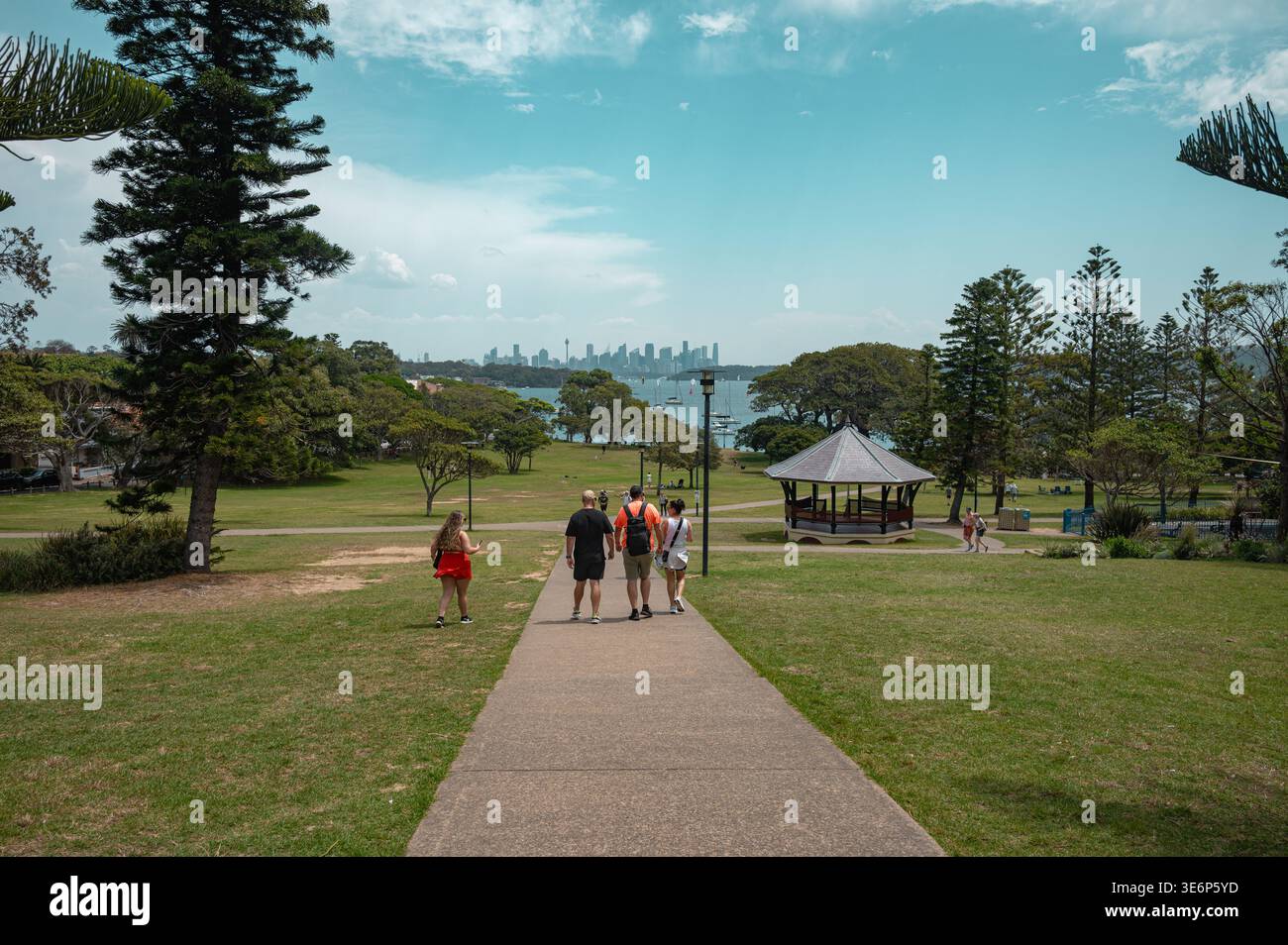 Persone che camminano su un sentiero nel Robertson Park passando davanti a un gazebo con una vista lontana dello skyline della città di Sydney dall'altra parte dell'acqua. Foto Stock