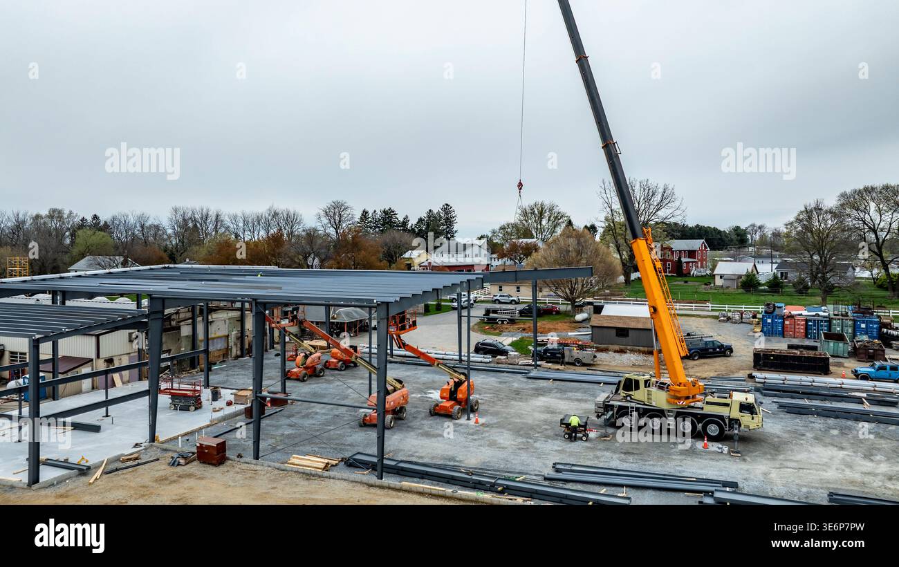 I lavoratori sono impegnati in un cantiere edile. Una grande gru solleva le travi in acciaio in posizione per un nuovo edificio. Sono presenti varie macchine e veicoli e alcune persone stanno guidando il lavoro. Foto Stock