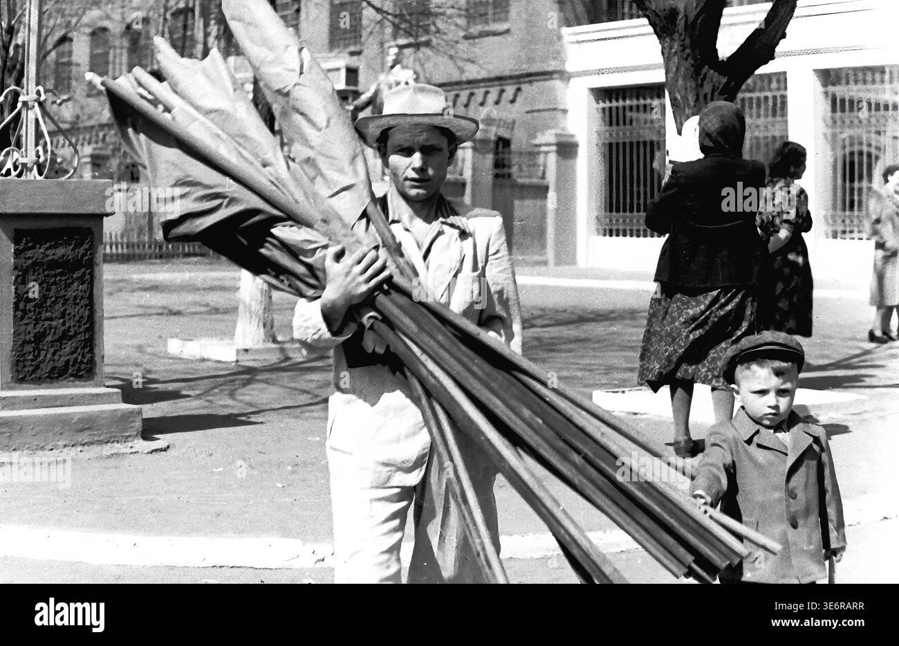 Sloviansk, 1950 anni. All'esterno dell'impianto di armatura e isolamento (AIZ). Un operaio sovietico dedicato che indossa un cappello sta trasportando un pesante fascio di lunghi pali di legno e striscioni di tessuto arrotolato in preparazione di una manifestazione pubblica festosa. Un ragazzino con un cappotto scuro si trova tranquillamente nelle vicinanze, sulla strada soleggiata. Questa autentica fotografia d'archivio in bianco e nero documenta perfettamente i preparativi attivi della comunità per le feste di massa, la vita urbana di strada quotidiana e l'autentico patrimonio culturale del pacifico Donbas. Foto Stock