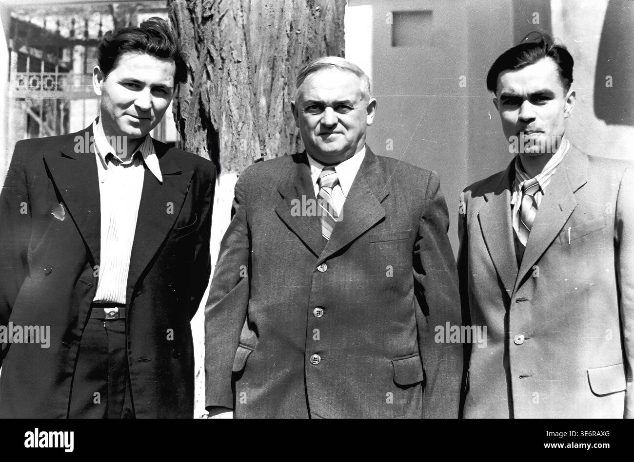 Sloviansk, 1950 anni. All'esterno dell'impianto di armatura e isolamento (AIZ). Tre fiduciosi uomini sovietici vestiti con abiti formali affilati posano insieme accanto a un tronco di albero grezzo nel cortile della fabbrica. I colleghi attendono con calma l'inizio di una manifestazione di massa festosa in una giornata molto luminosa e soleggiata. Questa fotografia d'archivio in bianco e nero altamente autentica mostra splendidamente la moda maschile della metà del secolo, le forti amicizie professionali, l'amministrazione della fabbrica e il ricco patrimonio culturale del pacifico Donbas. Foto Stock
