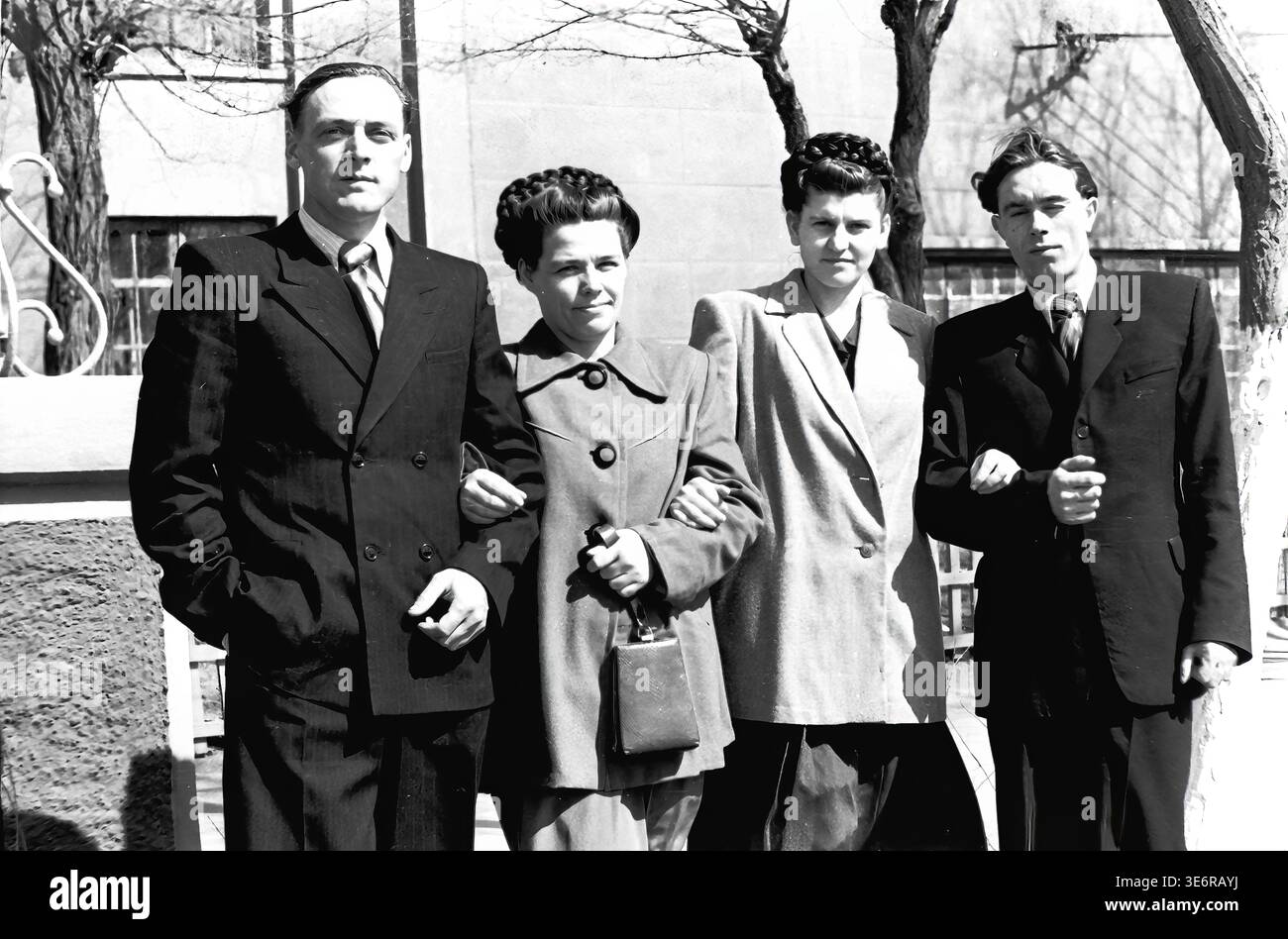 Sloviansk, 1950 anni. All'esterno dell'impianto di armatura e isolamento (AIZ). Quattro giovani colleghi della fabbrica sovietica si alzano con orgoglio al braccio, in posa per un ritratto di gruppo amichevole durante una luminosa giornata di sole prima di una festosa manifestazione di massa. Gli uomini indossano abiti scuri acuti e le donne sono vestite con eleganti cappotti della metà del secolo con bellissime acconciature intrecciate. Questa fotografia d'archivio in bianco e nero altamente autentica cattura perfettamente l'amicizia sul posto di lavoro, la cultura giovanile e il patrimonio della pace del Donbas. Foto Stock