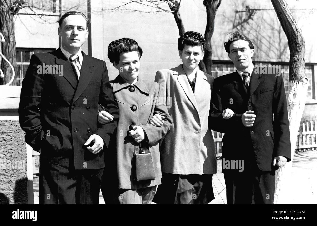 Sloviansk, 1950 anni. All'esterno dell'impianto di armatura e isolamento (AIZ). Quattro giovani colleghi della fabbrica sovietica si alzano con orgoglio al braccio, in posa per un ritratto di gruppo amichevole durante una luminosa giornata di sole prima di una festosa manifestazione di massa. Gli uomini indossano abiti scuri acuti e le donne sono vestite con eleganti cappotti della metà del secolo con bellissime acconciature intrecciate. Questa fotografia d'archivio in bianco e nero altamente autentica cattura perfettamente l'amicizia sul posto di lavoro, la cultura giovanile e il patrimonio della pace del Donbas. Foto Stock