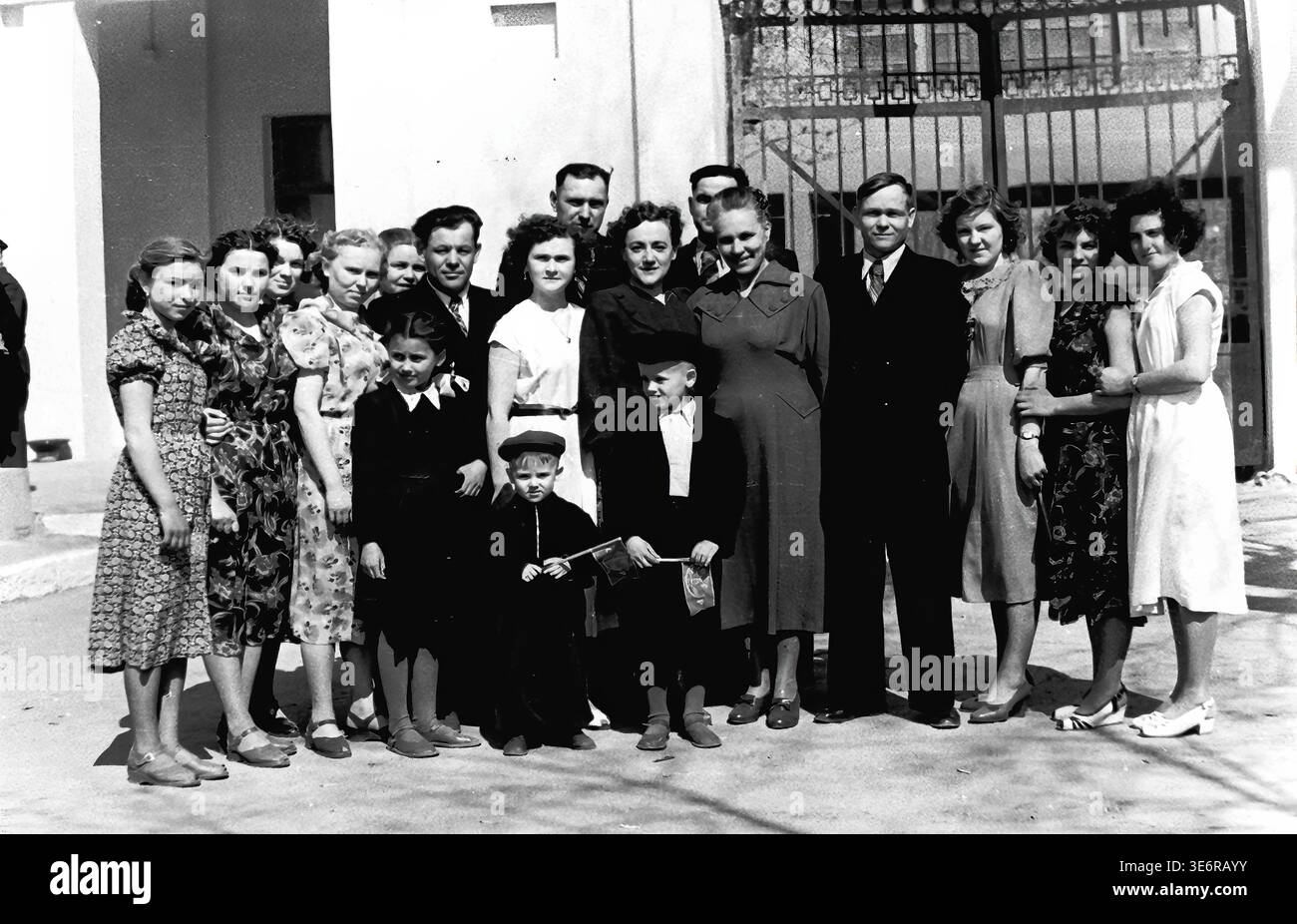 Sloviansk, 1950 anni. All'esterno dell'impianto di armatura e isolamento (AIZ). Un grande gruppo multigenerazionale di operai sovietici e le loro famiglie posa per un memorabile ritratto collettivo da un cancello di metallo decorativo prima di una festosa manifestazione di massa. I bambini sono felici di tenere piccole bandiere per le vacanze. Questa fotografia d'archivio in bianco e nero altamente autentica documenta brillantemente le riunioni della comunità della metà del secolo, la classica moda urbana, l'unità familiare e la storia culturale duratura del pacifico Donbas. Foto Stock