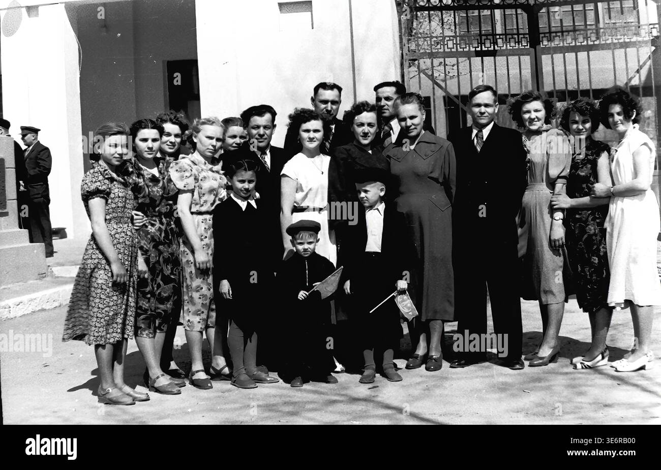 Sloviansk, 1950 anni. All'esterno dell'impianto di armatura e isolamento (AIZ). Un grande gruppo multigenerazionale di operai sovietici e le loro famiglie posa per un memorabile ritratto collettivo da un cancello di metallo decorativo prima di una festosa manifestazione di massa. I bambini sono felici di tenere piccole bandiere per le vacanze. Questa fotografia d'archivio in bianco e nero altamente autentica documenta brillantemente le riunioni della comunità della metà del secolo, la classica moda urbana, l'unità familiare e la storia culturale duratura del pacifico Donbas. Foto Stock