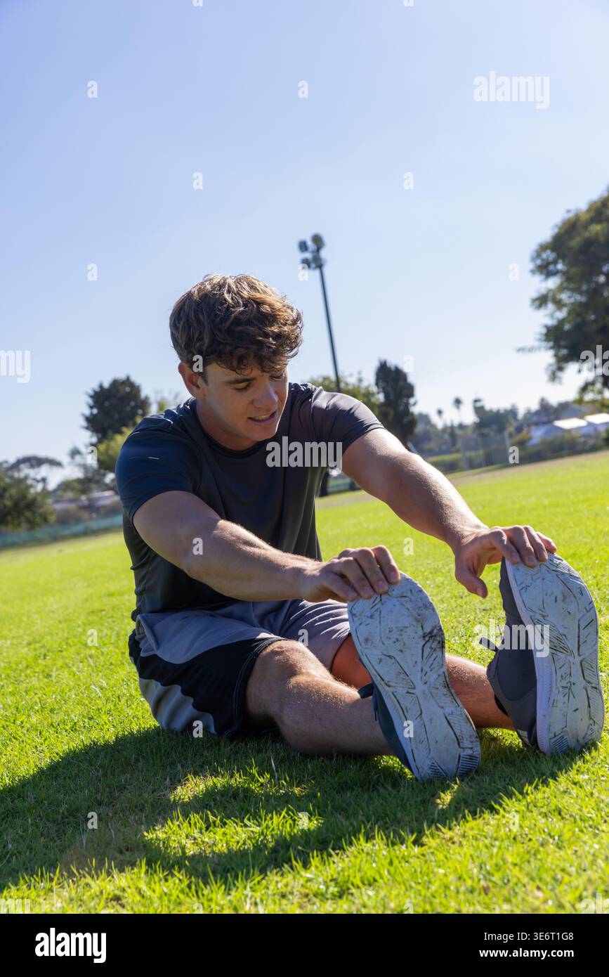 Uomo di 20 anni che allunga le gambe sul campo erboso, tiene in mano scarpe da ginnastica, indossa camicia e pantaloncini sportivi Foto Stock