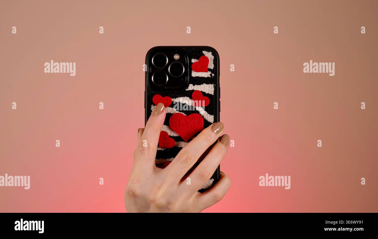 Una mano femminile tiene uno smartphone in una custodia con stampa zebrata e cuori rossi. Concetto di incontri online, amore digitale e saluti di vacanza moderni. Foto Stock