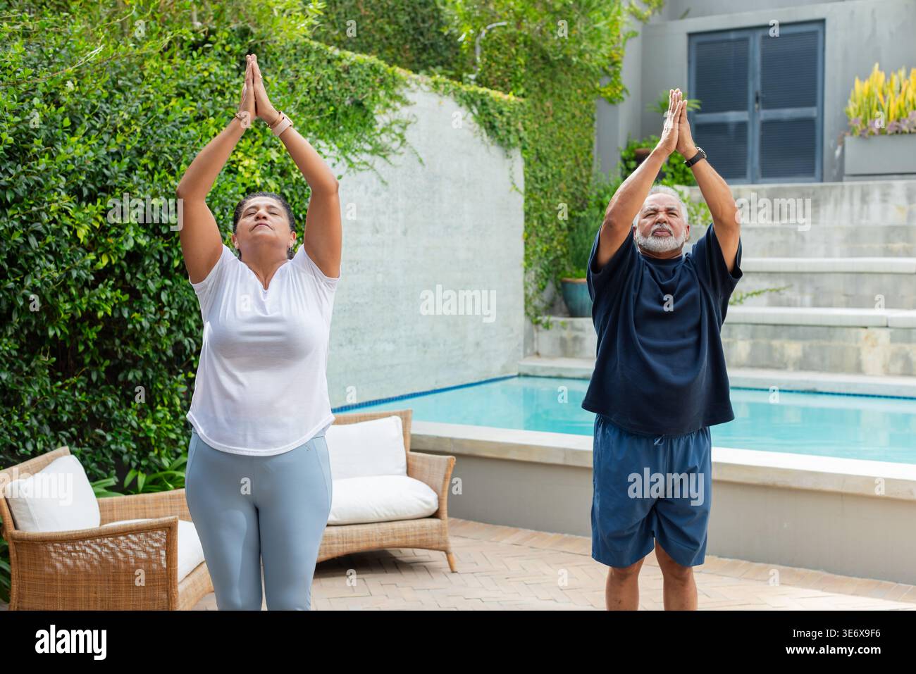 Coppie africane che fanno yoga sul patio piastrellato accanto alla piscina, indossano braccialetti e guardano Foto Stock