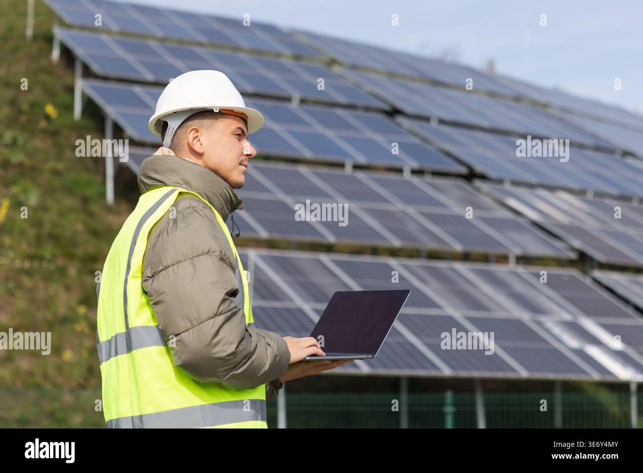 Un giovane ingegnere elettrico maschio ispeziona i sistemi a pannelli solari con un laptop in un ambiente a energia rinnovabile. Foto Stock