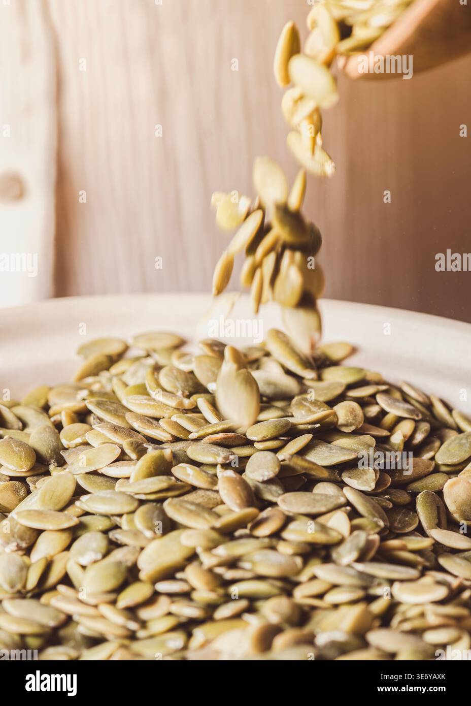 Semi di zucca che cadono da un cucchiaio di legno in un grande recipiente in ceramica bianca, calde tonalità dorate-beige, luce naturale soffusa, persona sullo sfondo, Co Foto Stock