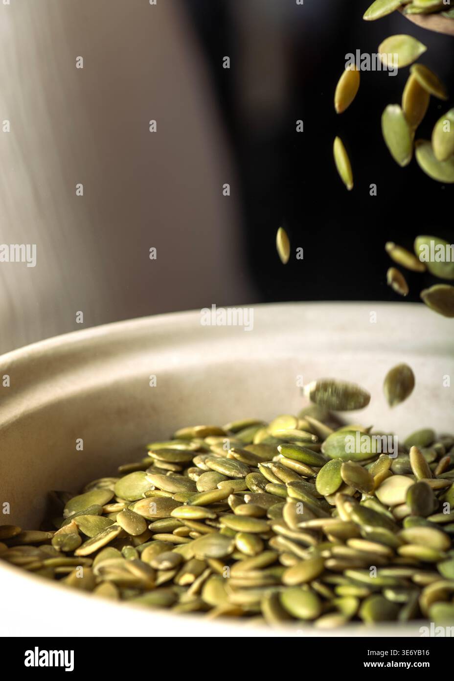 Semi verdi di zucca (pepitas) che cadono a mezz'aria in una ciotola di ceramica color crema, sfondo scuro drammatico, sfocatura del movimento, calda illuminazione laterale, rustica , copia spac Foto Stock
