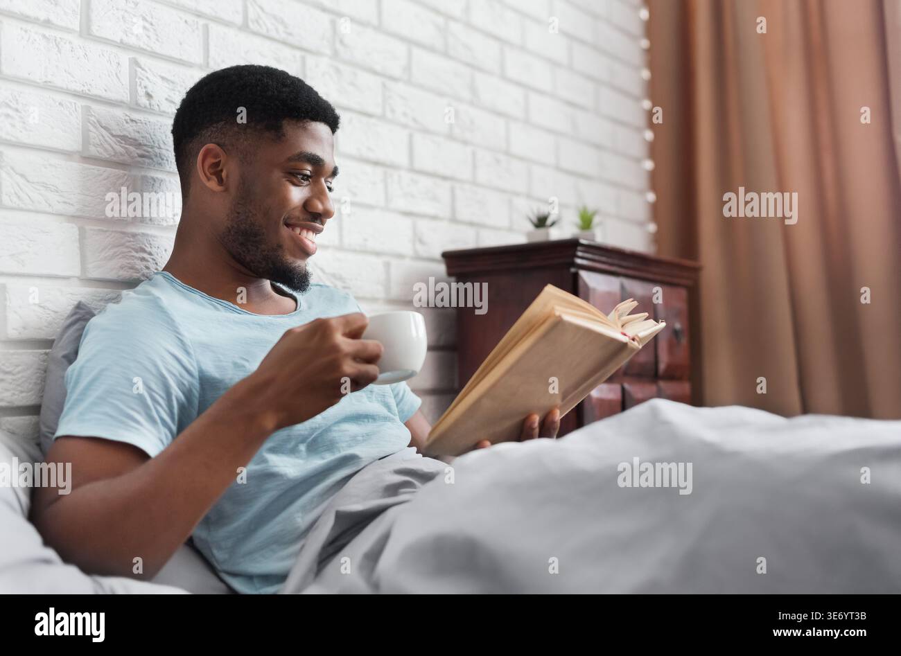 Uomo nero che beve caffè e legge un libro a letto Foto Stock