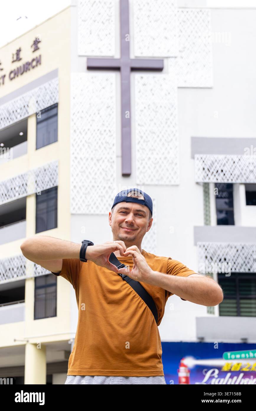 Un uomo sorridente che crea la forma del cuore con le mani in piedi davanti al moderno edificio della chiesa con croce. Fede, amore e concetto di comunità religiosa in urba Foto Stock