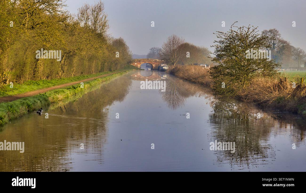 guardando lungo un canale, la nebbia mattutina. una barca stretta è ormeggiata in lontananza con un vecchio ponte in mattoni dietro di essa. i riflessi sono nell'acqua Foto Stock