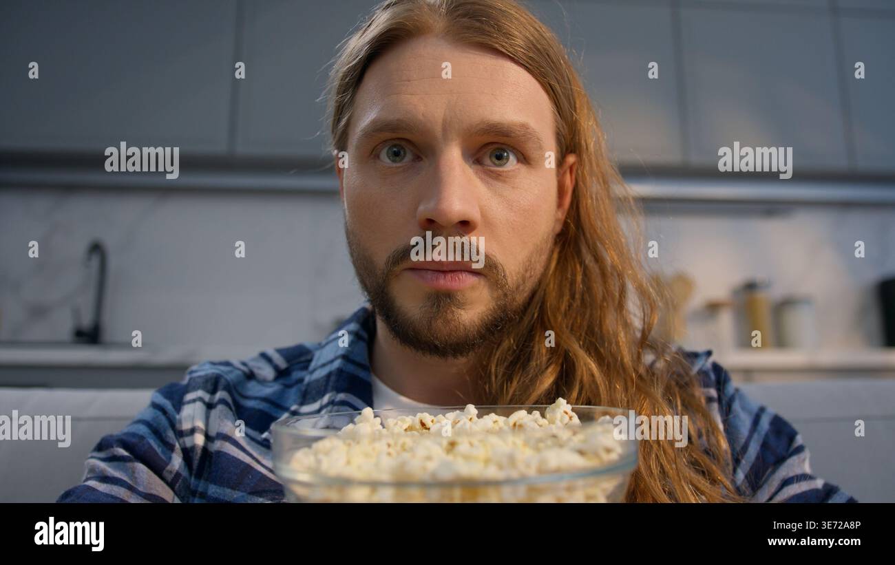 Un uomo caucasico concentrato che mangia popcorn guardando la TV a casa da vicino un ragazzo maschio che mangia gustosi snack di mais ossessionato dalla televisione di film Foto Stock