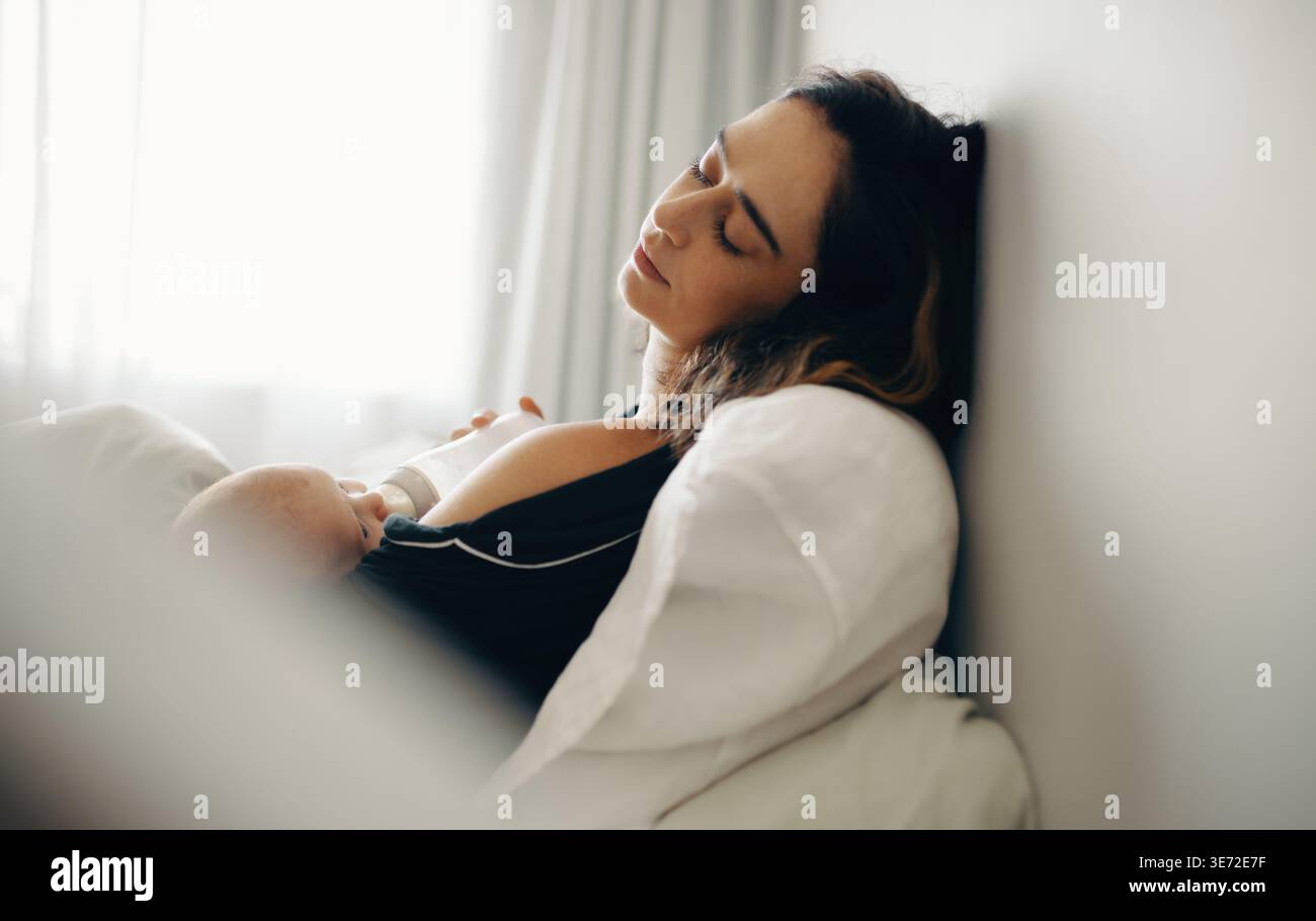 Una madre rilassata si appoggia su un cuscino mentre allatta il neonato con il biberon, catturando un momento di calma, cura e tranquillità Foto Stock