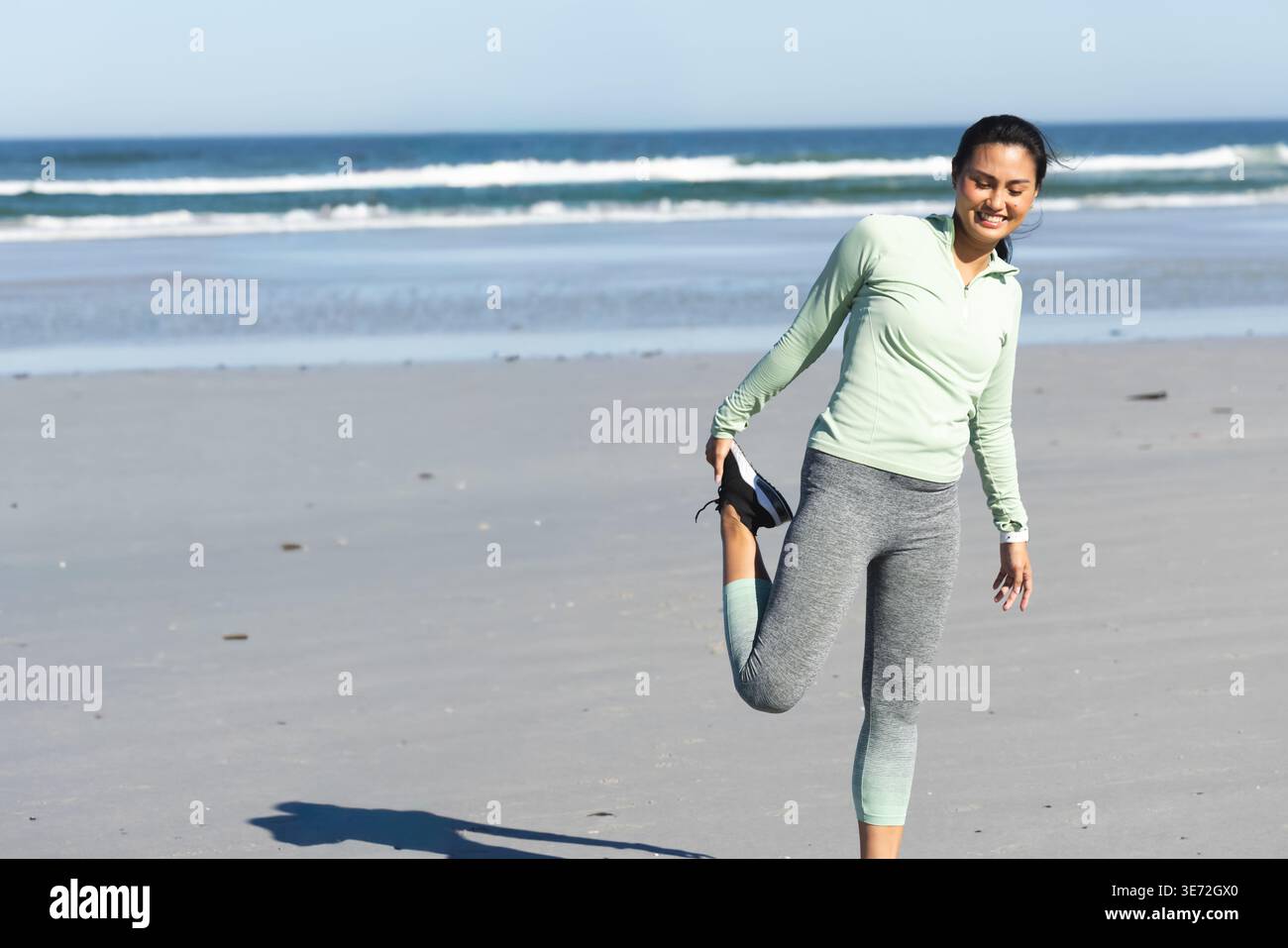 Donna asiatica che indossa leggings grigi con top verde che si estende su un quad sulla spiaggia sabbiosa che regge scarpe, copia spazio. Atletica, abbigliamento sportivo, outdoor, mare, onda Foto Stock