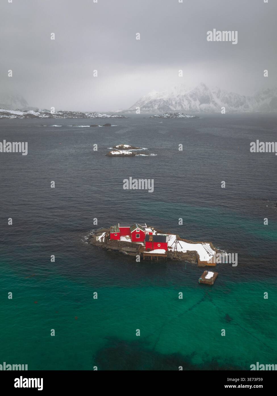 Vista aerea degli edifici rossi su un'isola rocciosa in contrasto con il mare blu profondo e il paesaggio innevato, Svolvær, Nordland, Norvegia. Foto Stock