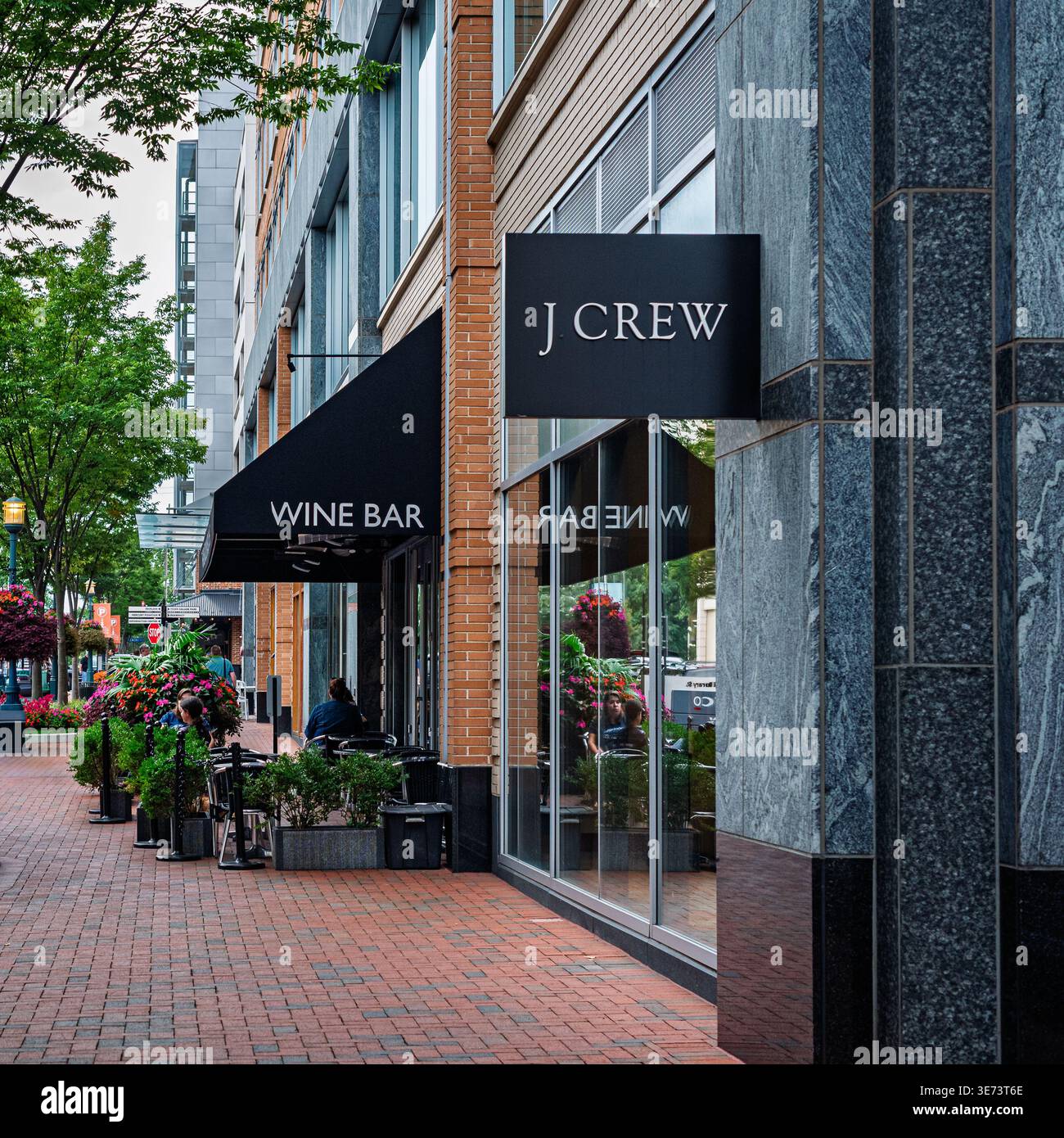 Un elegante paesaggio urbano caratterizzato da un punto vendita J.Crew accanto a un'area salotto wine bar all'aperto. La scena include marciapiedi in mattoni, moder Foto Stock