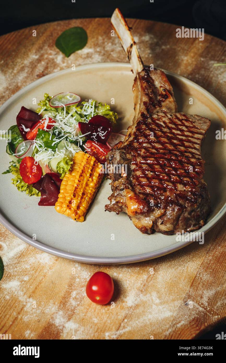 Bistecca gourmet di ribeye Tomahawk alla griglia con segni di ciccioli servita con insalata di mais fresco e pomodori ciliegini su un piatto di ceramica. Foto Stock
