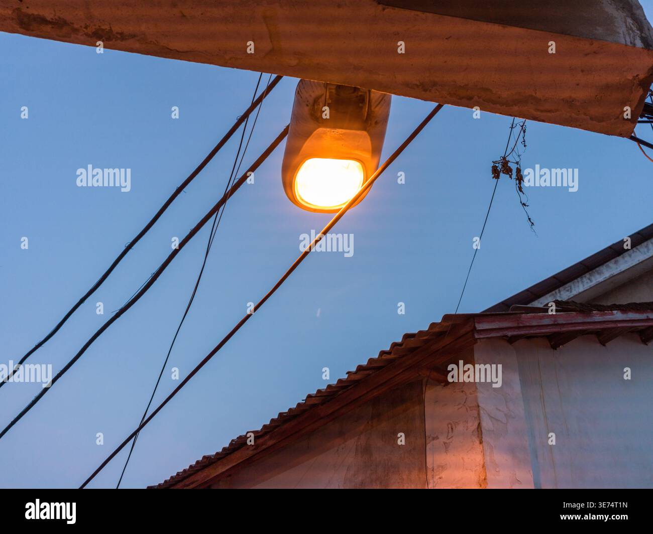 Un lampione arancione incandescente fissato a una struttura in legno illumina la scena lungo i cavi elettrici aggrovigliati e il bordo del tetto durante il twili profondo Foto Stock