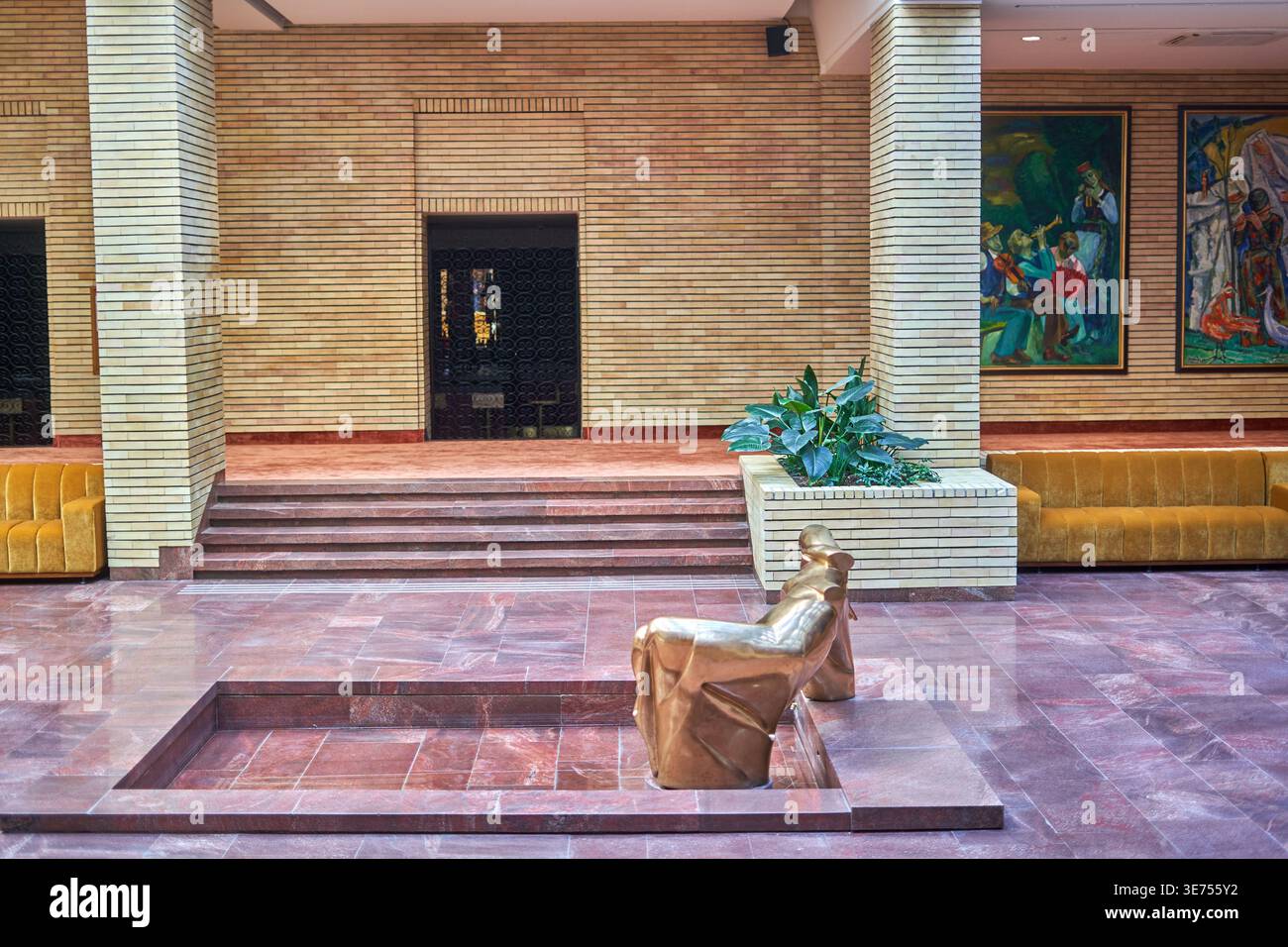 Un ampio atrio o lobby interno con pavimenti in marmo rosso lucido e pareti orizzontali in mattoni marrone chiaro. In primo piano, un'astratta in bronzo Foto Stock