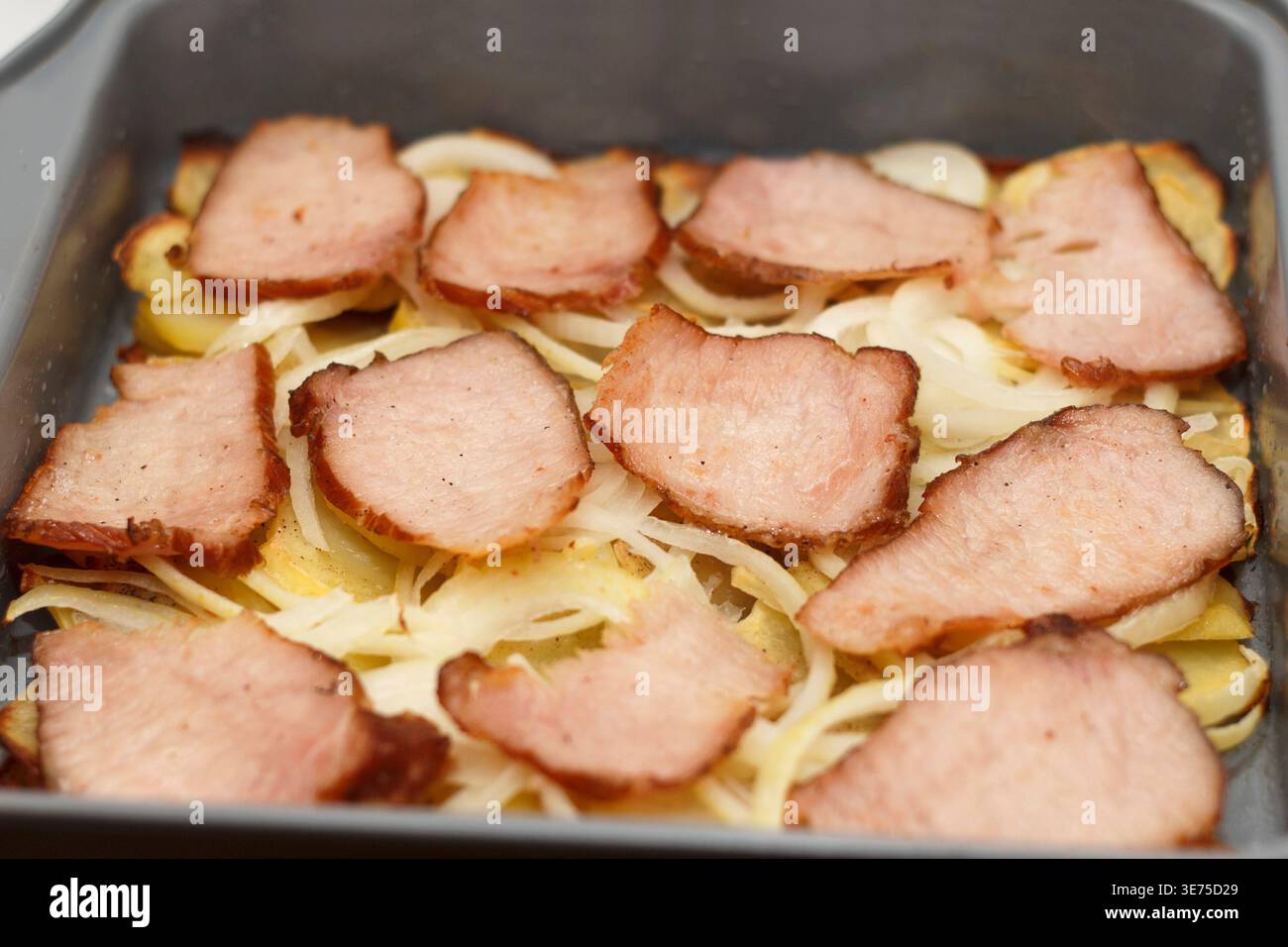 Primo piano di un piatto con carne affumicata affettata su un letto di cipolle e patate. Il cibo è disposto in modo ordinato in una teglia da forno Foto Stock