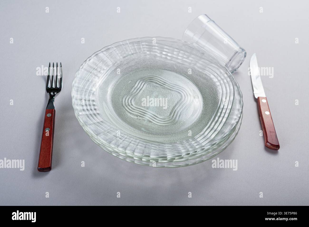 Configurazione minimalista con pila di piastre trasparenti, forchetta in metallo, coltello da cucina con manico in legno e bicchiere trasparente. Sfondo neutro, luce soffusa A. Foto Stock