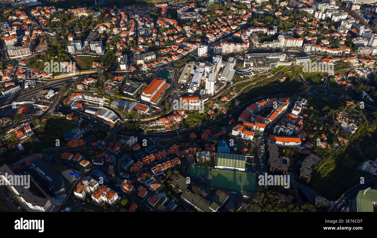 Vista aerea di Funchal, Madeira, incentrata su un grande ospedale con eliporto, strutture moderne, strade tortuose, terrazze, rotatorie, e pocket park Foto Stock
