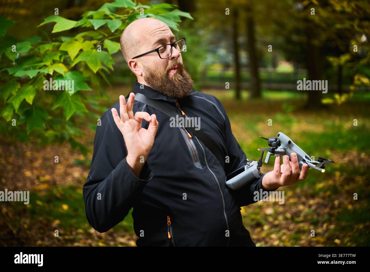 L'uomo fa gesti il segno OK tenendo il drone e il controller. L'uomo barbuto soddisfatto della configurazione nel parco, esprimendo approvazione e fiducia, concetto di successo, facilità d'uso e tecnologia Foto Stock