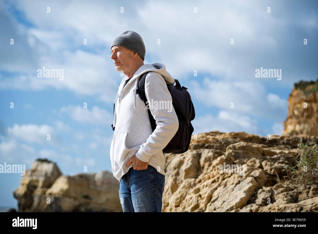 uomo che indossa un cappello di lana e una felpa con cappuccio in piedi sulle rocce con uno zaino, che guarda al mare Foto Stock