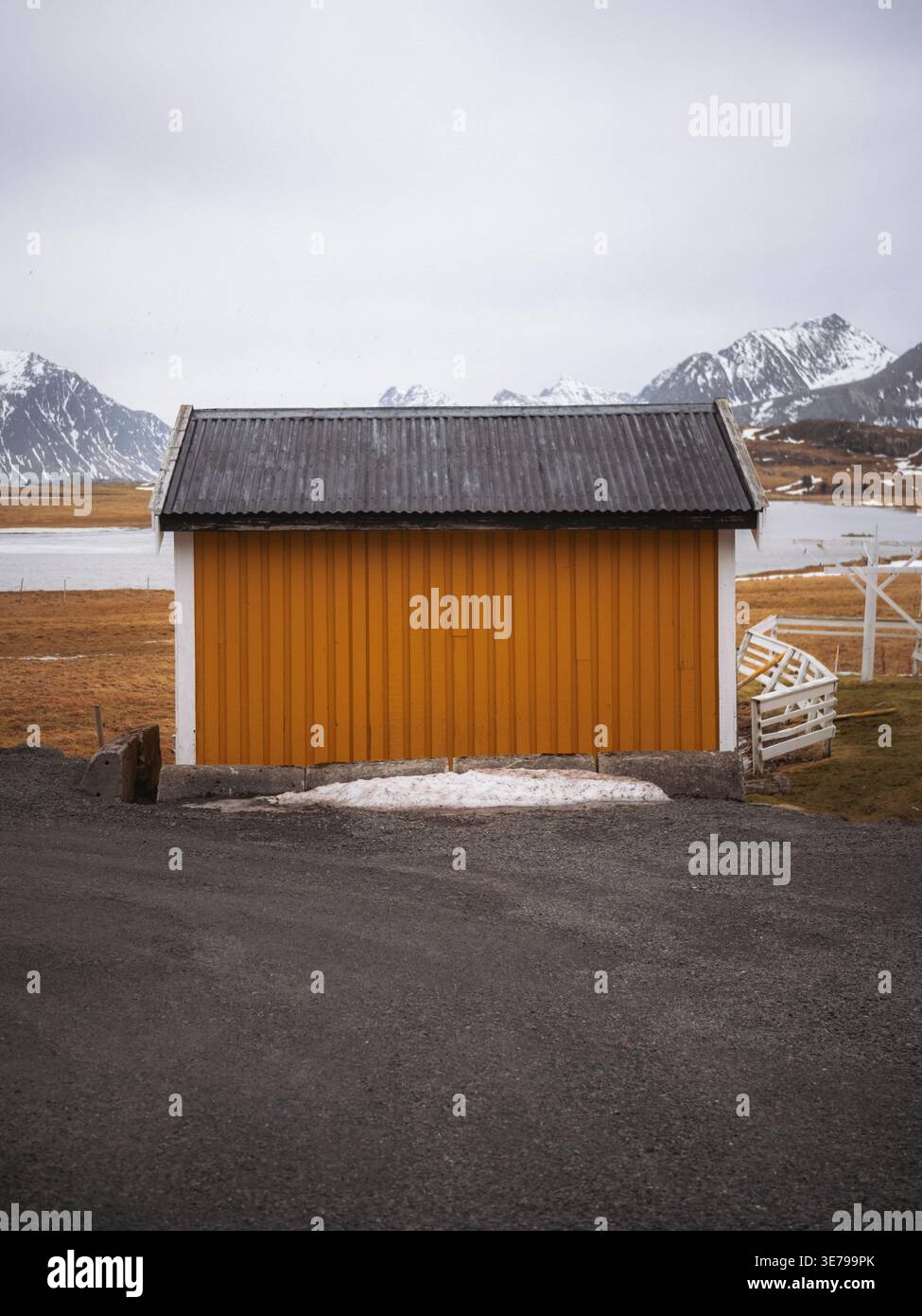 La vista di un edificio giallo senape con un tetto scuro si distingue contro le montagne innevate e i campi bruni, un netto contrasto nel tranquillo paesaggio Foto Stock