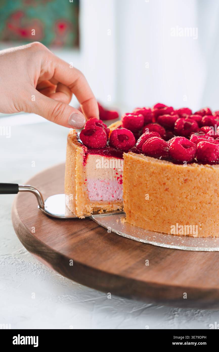 La mano prende una fetta di torta ai lamponi da un bordo di legno con frutti di bosco freschi in una cucina luminosa, momento del dessert, cottura fatta in casa Foto Stock