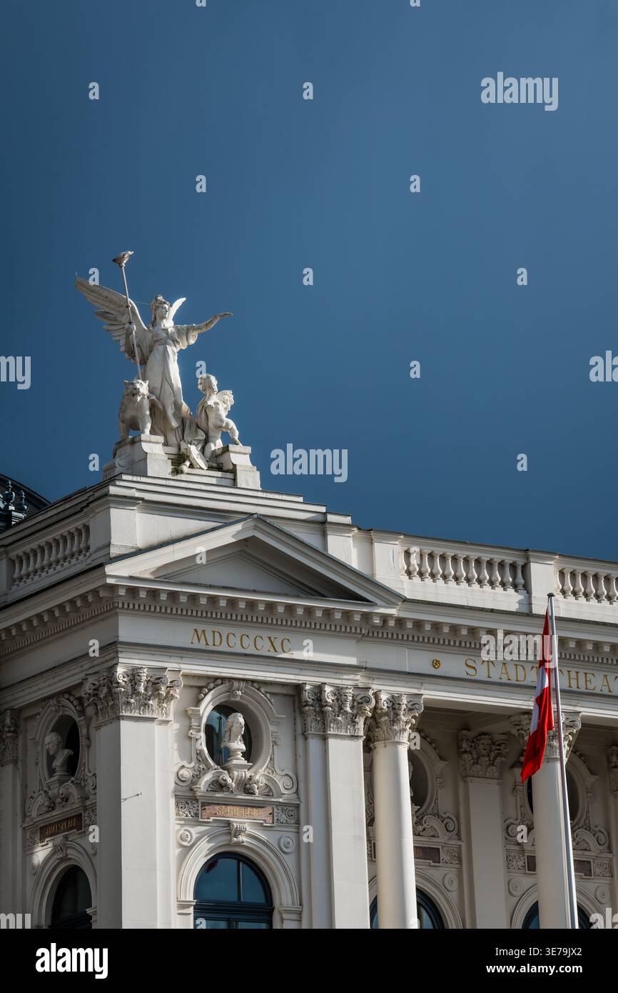 Zurigo, Svizzera - 2 aprile 2023: Vista della Schauspielhaus, la sua elegante facciata adornata con sculture sotto un cielo blu profondo, una bandiera svizzera delicatamente Foto Stock