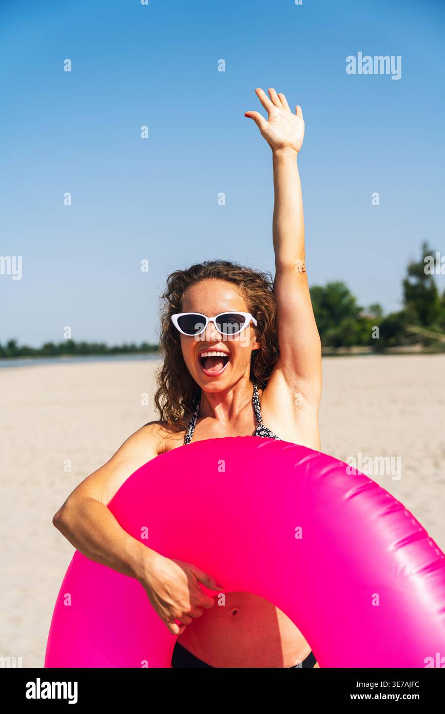 Donna allegra sulla spiaggia con anello gonfiabile rosa che ondeggia la mano, atmosfera di vacanza. Foto Stock