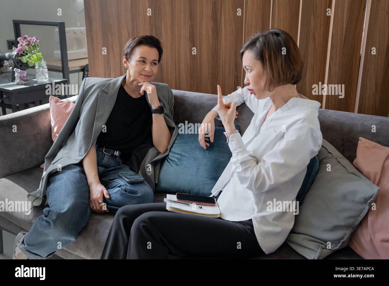Donne d'affari che conversano con i gesti delle mani sedute sul divano in un elegante interno dell'ufficio. Conversazione, gesti, ufficio, lavoro di squadra, affari Foto Stock
