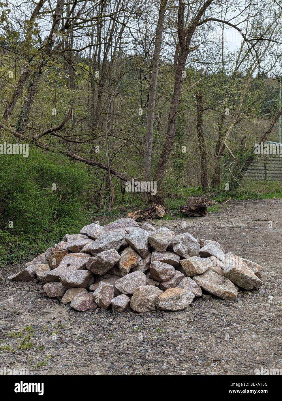 Una fotografia verticale di un mucchio di pietre di granito naturale grigie e marroni sedute su un sentiero sterrato con uno sfondo di alberi primaverili in erba e un fiume Foto Stock